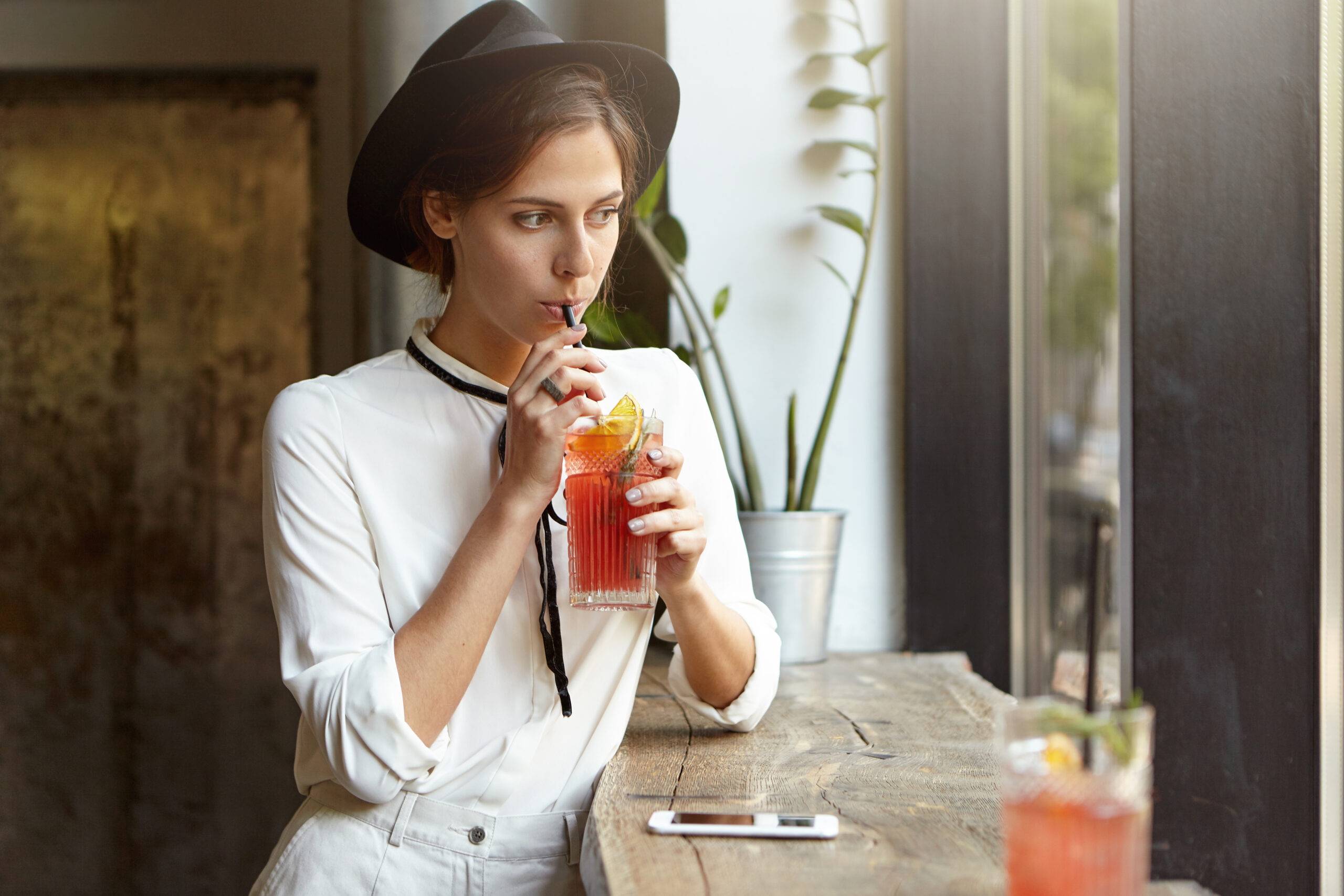 Attractive cute Caucasian girl in white jeans, blouse and black headwear sipping cold citrus juice or lemonade, sitting at cafe table by the window alone, looking outside with thoughtful expression